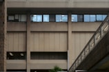 An urban building facade with a large, windowed concrete exterior and a connecting pedestrian bridge featuring metal railings. The windows reflect interior lighting, and a small portion of greenery is visible at the base.