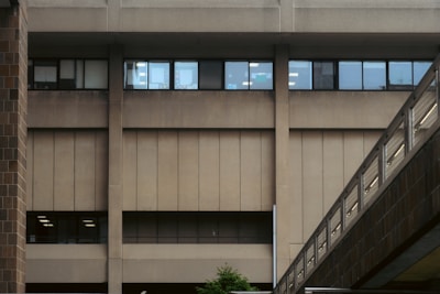 An urban building facade with a large, windowed concrete exterior and a connecting pedestrian bridge featuring metal railings. The windows reflect interior lighting, and a small portion of greenery is visible at the base.