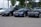 A trio of sleek BMW cars are parked on a paved area, with a backdrop of green trees and a billboard. The cars are positioned in a row, showcasing different shades of black and blue. The environment suggests a car dealership or a parking area.