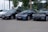 A trio of sleek BMW cars are parked on a paved area, with a backdrop of green trees and a billboard. The cars are positioned in a row, showcasing different shades of black and blue. The environment suggests a car dealership or a parking area.