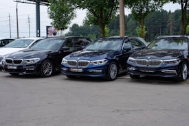 A trio of sleek BMW cars are parked on a paved area, with a backdrop of green trees and a billboard. The cars are positioned in a row, showcasing different shades of black and blue. The environment suggests a car dealership or a parking area.