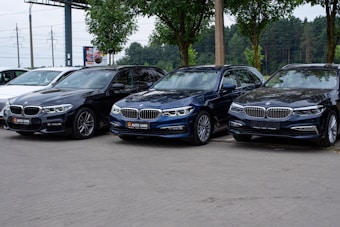 A trio of sleek BMW cars are parked on a paved area, with a backdrop of green trees and a billboard. The cars are positioned in a row, showcasing different shades of black and blue. The environment suggests a car dealership or a parking area.