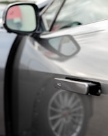 A close-up view of a car door handle that is sleek, modern, and partially extended. The focus is on the handle, with the side mirror and part of the car's body visible in the background. The design appears to be minimalist and possibly high-tech.