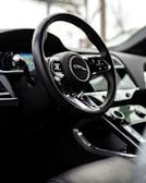 A close-up view of a car's interior, focusing on the steering wheel with a luxury logo. The dashboard features modern controls and a digital display. The surrounding environment suggests a sleek, high-quality design.