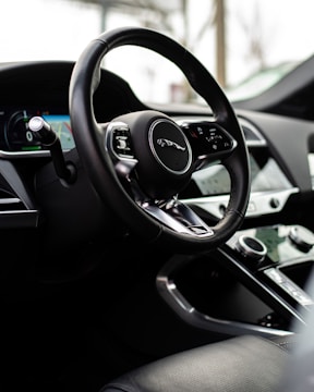 A close-up view of a car's interior, focusing on the steering wheel with a luxury logo. The dashboard features modern controls and a digital display. The surrounding environment suggests a sleek, high-quality design.