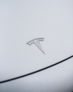 A close-up view of a metallic emblem of the Tesla logo, positioned on the smooth surface of a vehicle. The car's surface has small water droplets, indicating it might have rained recently.