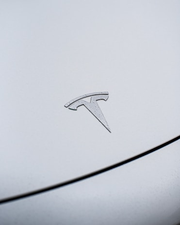 A close-up view of a metallic emblem of the Tesla logo, positioned on the smooth surface of a vehicle. The car's surface has small water droplets, indicating it might have rained recently.