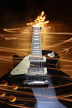 A black electric guitar with a white pickguard is positioned horizontally, surrounded by dynamic, swirling light trails that create a sense of movement and energy.