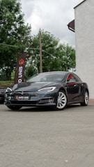 A sleek black car is parked on a paved surface. The vehicle is a Tesla, identifiable by its logo on the front. Nearby, a tall flag with the words 'AUTO UNIS' stands against a backdrop of green trees. Part of a building with a light-colored wall is visible on the right.