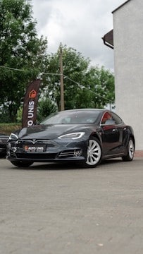 A sleek black car is parked on a paved surface. The vehicle is a Tesla, identifiable by its logo on the front. Nearby, a tall flag with the words 'AUTO UNIS' stands against a backdrop of green trees. Part of a building with a light-colored wall is visible on the right.