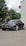 A sleek black car is parked on a paved surface. The vehicle is a Tesla, identifiable by its logo on the front. Nearby, a tall flag with the words 'AUTO UNIS' stands against a backdrop of green trees. Part of a building with a light-colored wall is visible on the right.