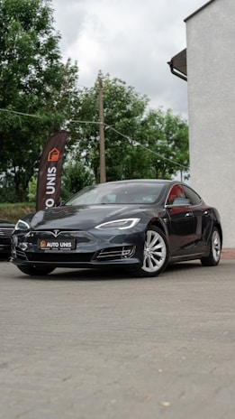 A sleek black car is parked on a paved surface. The vehicle is a Tesla, identifiable by its logo on the front. Nearby, a tall flag with the words 'AUTO UNIS' stands against a backdrop of green trees. Part of a building with a light-colored wall is visible on the right.