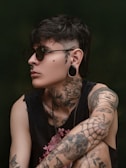 A person with dark hair and sunglasses is adorned with various tattoos including roses, a woman's face, and geometric patterns. They also have a nose piercing, earrings, and wear a sleeveless black shirt. The background is a muted green, giving a moody atmosphere.