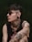 A person with dark hair and sunglasses is adorned with various tattoos including roses, a woman's face, and geometric patterns. They also have a nose piercing, earrings, and wear a sleeveless black shirt. The background is a muted green, giving a moody atmosphere.