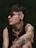 A person with dark hair and sunglasses is adorned with various tattoos including roses, a woman's face, and geometric patterns. They also have a nose piercing, earrings, and wear a sleeveless black shirt. The background is a muted green, giving a moody atmosphere.