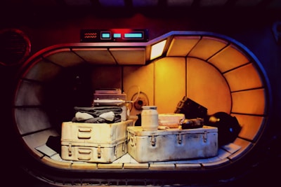 A futuristic storage compartment with a metallic interior contains various items. The items include two large, weathered storage containers, a pile of folded clothes, a jar, and other miscellaneous gear. Soft warm lighting illuminates the space, casting shadows and enhancing the metallic and industrial feel.