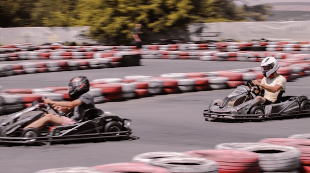 Two people are racing go-karts on a curving track surrounded by stacked tires painted in red, white, and black. Both individuals are wearing helmets as they speed around a bend. The background features trees and a blurred figure watching the race.