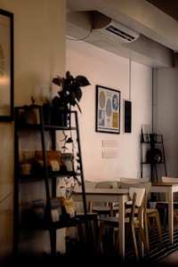 A cozy interior scene featuring a small coffee shop or café. Wooden tables and chairs are neatly arranged, with decorative plants and framed artwork on the walls. Shelves showcase various small items, possibly products for sale. The lighting is warm and intimate, creating an inviting atmosphere.