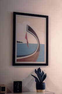 A framed geometric artwork features a stylized sailboat against a light background, with a red sail and a blue sky. Below the frame, there is a dark blue potted plant with pointed leaves set on a surface beside a black clock showing approximately 3:13.