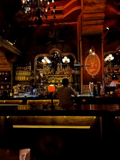 A dimly lit bar with a cozy and warm ambiance. The decor includes an ornate chandelier hanging from the ceiling, a well-stocked bar with various bottles of alcohol, and soft lighting creating an inviting atmosphere. A person is sitting at the bar, facing away, wearing a striped shirt. Decorative elements and vintage signs are visible on the walls, contributing to the rustic charm.