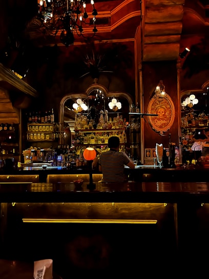 A dimly lit bar with a cozy and warm ambiance. The decor includes an ornate chandelier hanging from the ceiling, a well-stocked bar with various bottles of alcohol, and soft lighting creating an inviting atmosphere. A person is sitting at the bar, facing away, wearing a striped shirt. Decorative elements and vintage signs are visible on the walls, contributing to the rustic charm.