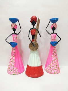 Three artistic sculptures depicting stylized figures, each adorned with traditional headgear and long dresses. The two figures on the sides carry blue baskets on their heads while the central figure holds a circular object and some sticks. The dresses feature intricate floral patterns.