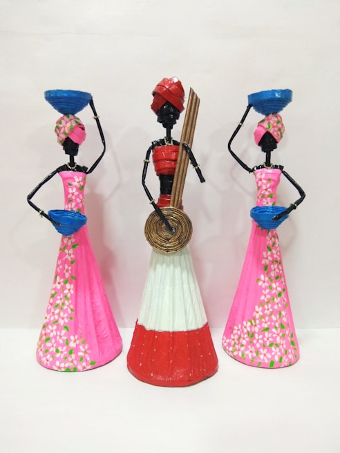 Three artistic sculptures depicting stylized figures, each adorned with traditional headgear and long dresses. The two figures on the sides carry blue baskets on their heads while the central figure holds a circular object and some sticks. The dresses feature intricate floral patterns.
