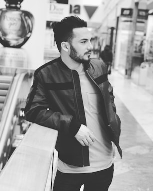 A man is leaning casually on a railing in a shopping mall. He is wearing a jacket and a T-shirt, with well-groomed hair and a beard. The setting includes escalators and various stores, and a large perfume bottle advertisement is visible in the background.