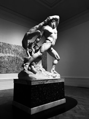 A powerful marble sculpture depicting a muscular figure with dynamic posture, possibly engaged in an intense action. The room is softly lit, enhancing the dramatic shadows and highlighting the intricate details of the statue. The setting appears to be an art gallery with spacious, minimal decor.