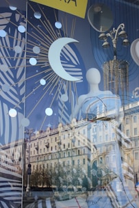 A reflective storefront display showcases celestial-themed decor with a prominent crescent moon and starburst patterns. The mannequin, partially visible, is adorned with the word 'dream'. Visible is a reflection of an ornate street lamp and a stately building, adding architectural interest to the scene.