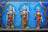 Three intricately designed statues adorned with vibrant garlands and traditional attire are set against a backdrop of blue with embellished arch designs. The figures appear to be deities holding staffs, and the images are richly decorated with floral offerings, including marigolds.