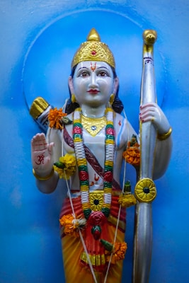 A colorful statue of a deity adorned with garlands and flowers. The deity is holding a bow and is dressed in vibrant traditional attire, including a golden headdress and jewelry. The statue has intricate detailing and is set against a light blue background.