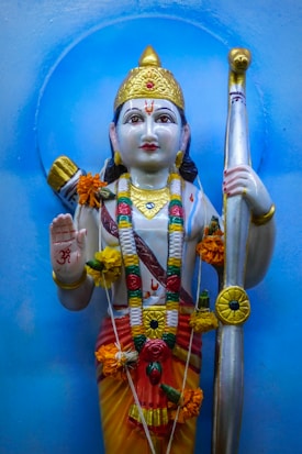 A colorful statue of a deity adorned with garlands and flowers. The deity is holding a bow and is dressed in vibrant traditional attire, including a golden headdress and jewelry. The statue has intricate detailing and is set against a light blue background.