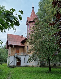 A rustic house with a tall, pointed tower and a red roof is surrounded by lush greenery. An ornate metal awning covers the entrance, and a tree with clusters of red berries stands to the right, adding to the quaint atmosphere.