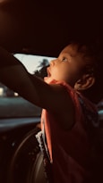 A young child with curly hair and a focused expression reaches upward inside a car, highlighted by soft, warm lighting. The child is wearing a red garment, and the interior of the car is dimly lit. Outside, faint traces of a landscape are visible through the windows.