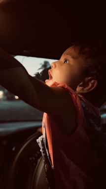A young child with curly hair and a focused expression reaches upward inside a car, highlighted by soft, warm lighting. The child is wearing a red garment, and the interior of the car is dimly lit. Outside, faint traces of a landscape are visible through the windows.