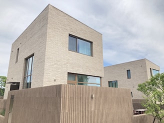 Two modern, minimalistic houses with light gray brick exteriors. The structures are geometric and their design appears sleek and contemporary. Surrounding the houses is a fence made of vertical slats, with some greenery visible in the foreground.