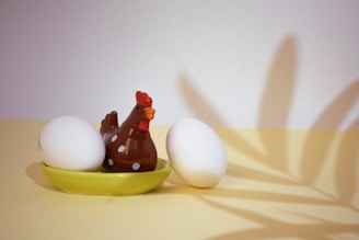 A ceramic chicken figure with white polka dots is placed on a yellow dish alongside two white eggs. A shadow, resembling leaves, is cast on the wall behind, adding a decorative element to the composition.