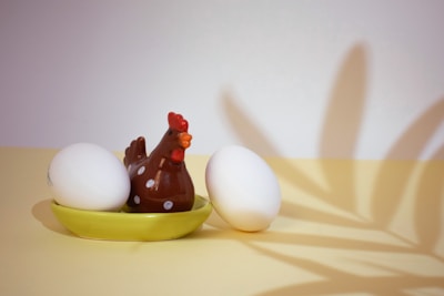 A ceramic chicken figure with white polka dots is placed on a yellow dish alongside two white eggs. A shadow, resembling leaves, is cast on the wall behind, adding a decorative element to the composition.