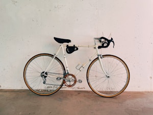 A vintage-style road bicycle is positioned against a plain white wall. The bicycle features drop handlebars, a black seat, and a tan leather saddle bag. The tires have a gum wall finish, and the frame is painted a light cream color with contrasting metallic components. The whole setup rests on a concrete floor.