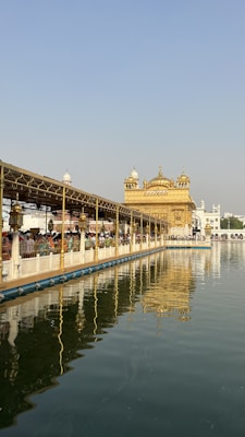 A stunning golden temple sits beside a serene body of water, reflecting its intricate architecture in the calm surface. The area is bustling with numerous people walking alongside the edge, under a long covered pathway. The sky is clear, accentuating the grandeur of the temple's gilded hues.
