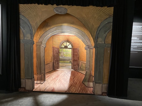 An artistic depiction of an arched hallway with columns leading to a door. The artwork features a warm color palette with shades of orange and brown, creating an inviting atmosphere. Beyond the open door, a bright landscape is visible through a circular window.