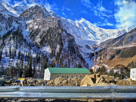 Majestic snow-covered mountains dominate the landscape, with rugged peaks and lush forests down the slopes. A vibrant blue sky with scattered clouds adds to the breathtaking scenery. Below, modest buildings with green roofs sit amidst rocky terrain.