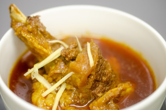 A white bowl filled with a rich, red curry containing a cooked meat piece garnished with thin slices of ginger. The dish appears hearty and aromatic.