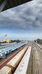 A large railway station with multiple platforms and tracks, featuring several trains lined up under a partly cloudy sky. The Indian national flag is visible, and there are buildings and trees in the background. The scene is captured from an elevated angle, revealing the scale and extent of the station.