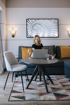 A woman sits at a modern round wooden table with a laptop in a stylish living room. The room features a gray sofa with yellow and gray pillows, a geometric patterned rug, and a decorative wall art piece. The lighting is soft, and a wine glass is placed on the table.