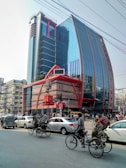 A tall modern building with a reflective glass facade and distinctive red accents is surrounded by a busy street scene. The street is bustling with various vehicles, including rickshaws, cars, and a unique three-wheeled vehicle. The urban environment includes other buildings, streetlights, and overhead cables.