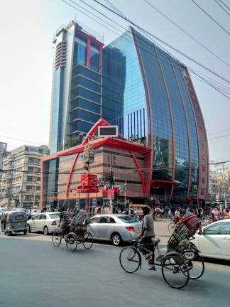 A tall modern building with a reflective glass facade and distinctive red accents is surrounded by a busy street scene. The street is bustling with various vehicles, including rickshaws, cars, and a unique three-wheeled vehicle. The urban environment includes other buildings, streetlights, and overhead cables.