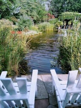A serene garden scene with a small pond bordered by lush vegetation and colorful flowers. Two white wooden chairs are positioned on a stone patio facing the pond, inviting relaxation. The background features various trees and shrubs with a pergola covered in greenery.