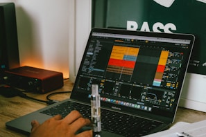 A person is using a MacBook Pro laptop with music production software open on the screen, displaying a project with multiple tracks and controls. The laptop is placed on a wooden desk beside a red audio interface. In the background, part of a poster with the word 'BASS' is visible.